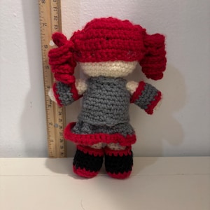 May include: A handmade crochet doll with a red hair, gray dress, and black shoes. The doll is approximately 6 inches tall and has a white face. The doll's dress has red trim, and the hair is styled in two pigtails.
