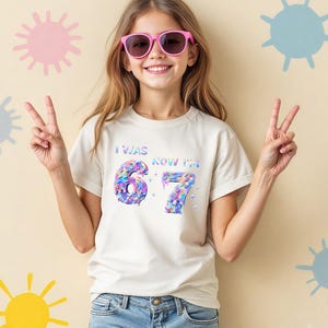 Peut inclure: Une jeune personne porte un t-shirt crème avec le texte "I WAS 6 NOW I'M 7" dans un design irisé coloré. La personne porte des lunettes de soleil roses et un jean bleu, faisant le signe de la paix avec les deux mains.