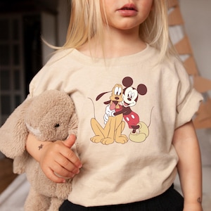 May include: A beige t-shirt featuring a graphic of Mickey Mouse and Pluto hugging. The shirt is worn by a child holding a stuffed bunny. The text "baby blossoms tees" is in the upper right corner.