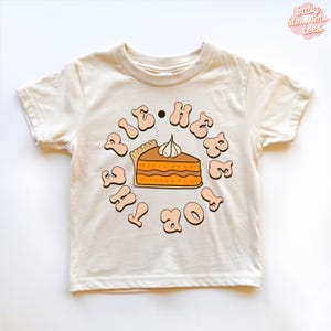 May include: A cream-coloured short-sleeve t-shirt featuring a central graphic of a slice of pumpkin pie topped with whipped cream. The text "HERE FOR THE PIE" is printed in a circular, wavy brown font surrounding the pie illustration.