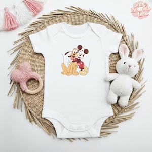 May include: White baby onesie featuring a graphic of Mickey Mouse and Pluto hugging. The onesie has short sleeves and a snap closure. The text "baby blossoms tees" is in the upper right corner. A pink heart-shaped rattle and a white stuffed bunny are also in the image.