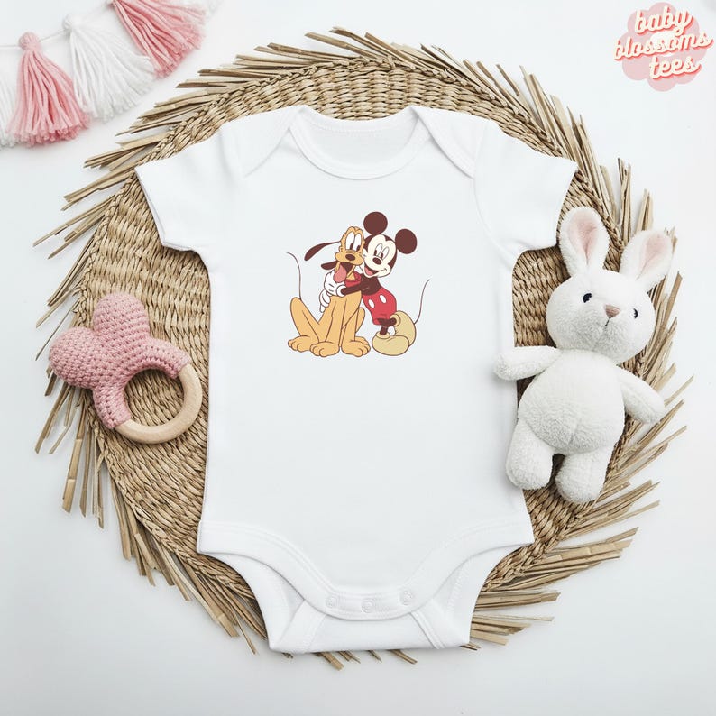 May include: White baby onesie featuring a graphic of Mickey Mouse and Pluto hugging. The onesie has short sleeves and a snap closure. The text "baby blossoms tees" is in the upper right corner. A pink heart-shaped rattle and a white stuffed bunny are also in the image.