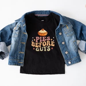 May include: A black t-shirt with the text "PIES BEFORE GUYS" in a retro font, featuring a pie graphic. A blue denim jacket is draped over the shirt. The shirt is likely for children.