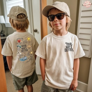 Op de afbeelding: Twee kinderen dragen crèmekleurige T-shirts. Eén shirt heeft de tekst "Your Way's Better" op de voorkant en een afbeelding op de achterkant. Het andere shirt heeft dezelfde tekst op de voorkant en een afbeelding op de achterkant. Beide kinderen dragen kaki petten.