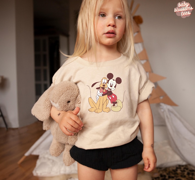 May include: A beige t-shirt featuring a graphic of Mickey Mouse and Pluto hugging. The shirt is worn by a child holding a stuffed bunny. The text "baby blossoms tees" is in the upper right corner.