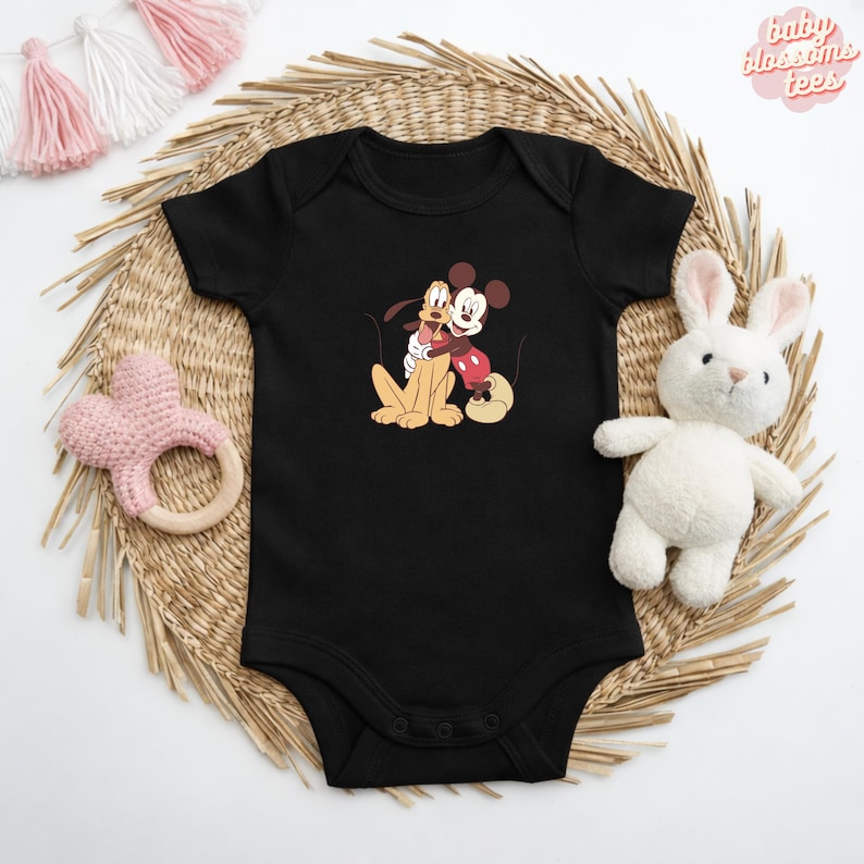 May include: Black baby bodysuit featuring a cartoon graphic of Mickey Mouse hugging Pluto. The bodysuit has short sleeves and snap closures. The text "baby blossoms tees" is in the upper right corner.