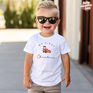 Pode incluir: Camiseta branca com o texto "MY FIRST Christmas" e um desenho de carro de corrida de desenho animado. A camiseta é usada por uma criança pequena usando óculos de sol pretos.