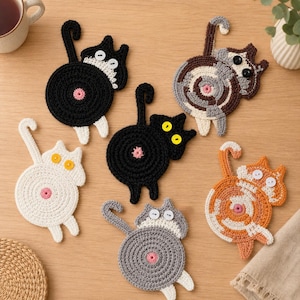 May include: A set of six crocheted cat coasters in various colours including black, white, grey, orange, and multi-coloured patterns. Each coaster features a circular body, a raised tail, two button eyes, and a small pink circular centre detail.