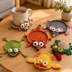 Peut inclure: Un ensemble de cinq dessous de verre ronds en crochet représentant des animaux : une souris beige sous une tasse blanche, une souris grise, un renard orange, une grenouille verte et un canard jaune. Chaque pièce a des yeux cousus et des membres.