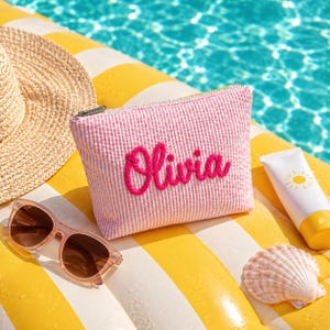 Könnte beinhalten: Eine rosafarbene und weiße Seersucker-Tasche mit Reißverschluss, auf der der Name Olivia in leuchtend pinkfarbenen Garnbuchstaben gestickt ist. Die Tasche hat eine rechteckige Form, einen Reißverschluss oben und eine strukturierte Stoffoberfläche.