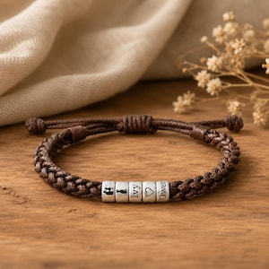 Könnte beinhalten: Ein braunes Armband aus geflochtener Kordel mit fünf silberfarbenen zylindrischen Perlen. Die Perlen sind mit einer Silhouette von zwei Personen, einer Katzensilhouette, dem Namen Ivy, einem Herzsymbol und dem Namen Owen in schwarzer Schrift graviert.