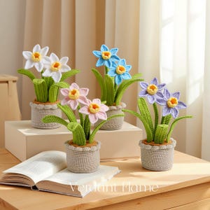 Könnte beinhalten: Gehäkelte Narzissenpflanzen in Weiß, Blau, Rosa und Lila, jede mit gelber Mitte und grünen Blättern. Die Pflanzen stehen in grauen, strukturierten Töpfen. Der Text "Verdant Flame" ist auf einem Buch zu sehen.
