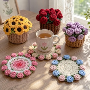 May include: Three crocheted flower pots featuring yellow sunflowers, red roses, and purple roses, alongside two round crocheted coasters with floral edges and a ceramic mug with a tulip illustration, all arranged on a wooden surface.