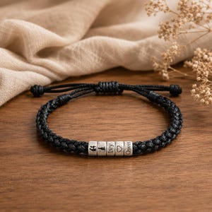 Puede incluir: Pulsera trenzada negra con cuentas plateadas. Las cuentas están grabadas con una corona, una pieza de ajedrez, un corazón y los nombres "Ivy" y "Owen". La pulsera es ajustable y está sobre una superficie de madera.