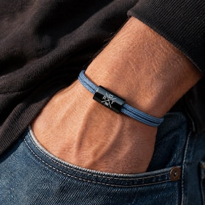 May include: A blue braided bracelet with a black clasp. The clasp features the letters "RXT", a heart symbol, and the number "96". The bracelet is worn on a wrist, with the person wearing a black top and blue jeans.