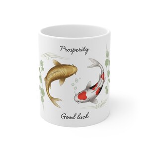 以下が含まれることがあります： 金と赤の鯉のイラストが描かれた白いセラミックマグカップ。マグカップには「Prosperity」と「Good luck」の文字が黒い筆記体で書かれています。緑の葉のアクセントと泡がデザインを完成させています。