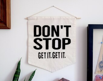 May include: A cream-coloured banner with the words "DON'T STOP" in large black letters, followed by "GET IT. GET IT." in smaller black letters. The banner is suspended from a wooden dowel with a string.