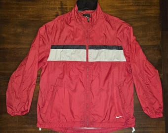 Puede incluir: Chaqueta cortavientos Nike roja con cuello negro y cremallera completa. La chaqueta presenta un diseño de rayas horizontales en negro y blanco en el pecho. Un pequeño logotipo blanco de Nike es visible en la parte inferior derecha.