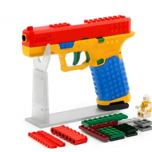 Puede incluir: Una pistola de juguete colorida construida con ladrillos de plástico entrelazados. La pistola presenta una parte superior roja, un cuerpo amarillo, un agarre azul y detalles verdes. Se incluyen ladrillos adicionales y una minifigura, todo exhibido en un soporte transparente.