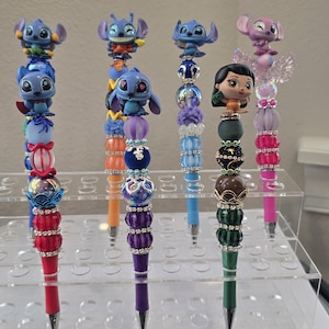 May include: A collection of decorative pens featuring character toppers. The pens have colorful beaded shafts and are topped with figures of Stitch, Angel, and Lilo & Stitch characters. The pens are displayed on a clear acrylic stand.