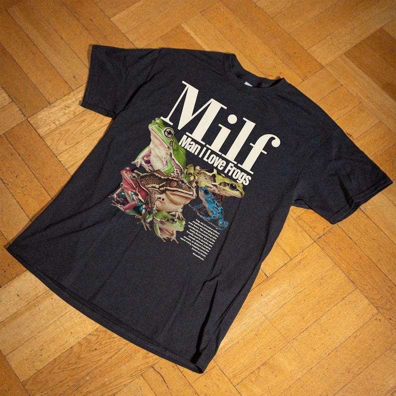 May include: Dark grey t-shirt with a colourful frog graphic and the text "Milf Man I Love Frogs" in white. The design features various frog species in shades of green, brown, pink, and blue.