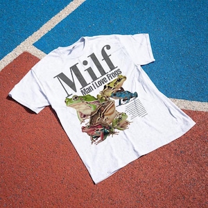 May include: White t-shirt with the text "Milf Man I Love Frogs" in black. The shirt features a graphic of several colourful frogs. The shirt is laying on a red and blue surface.