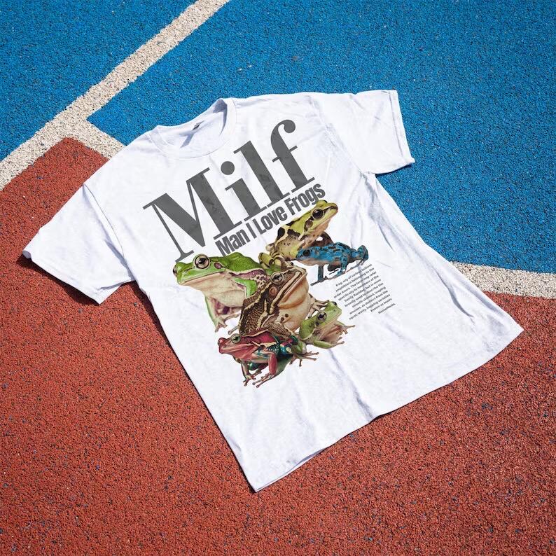 May include: White t-shirt with the text "Milf Man I Love Frogs" in black. The shirt features a graphic of several colourful frogs. The shirt is laying on a red and blue surface.