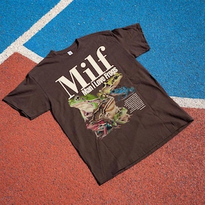 May include: A brown t-shirt with the text "Milf Man I Love Frogs" in white. The shirt features a graphic of several colourful frogs. The shirt is displayed on a red and blue surface.