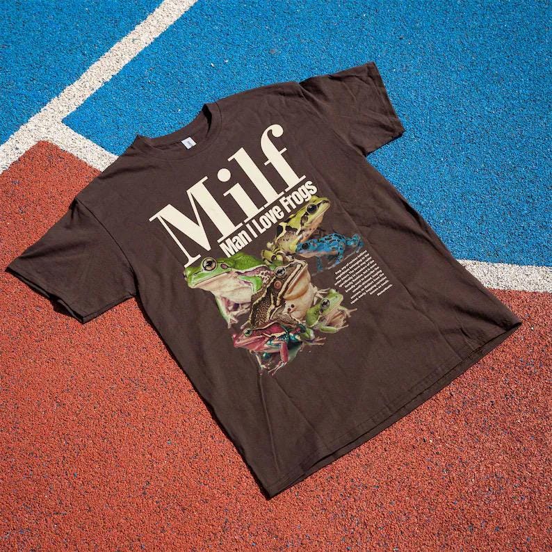 May include: A brown t-shirt with the text "Milf Man I Love Frogs" in white. The shirt features a graphic of several colourful frogs. The shirt is displayed on a red and blue surface.