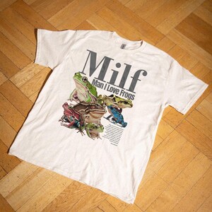 May include: White t-shirt with a graphic of various colourful frogs. The shirt displays the word "Milf" in large grey letters above the phrase "Man I Love Frogs". A small block of text is below the frogs.