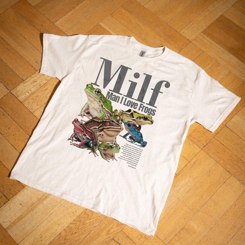 May include: White t-shirt with a graphic of various colourful frogs. The shirt displays the word "Milf" in large grey letters above the phrase "Man I Love Frogs". A small block of text is below the frogs.