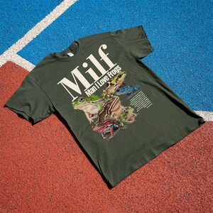 May include: Dark green t-shirt with the text "Milf" in large white letters, with "Man I Love Frogs" below. The shirt features a graphic of several colourful frogs. The shirt is lying on a red and blue surface.