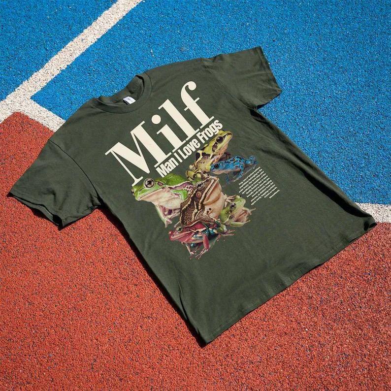 May include: Dark green t-shirt with the text "Milf" in large white letters, with "Man I Love Frogs" below. The shirt features a graphic of several colourful frogs. The shirt is lying on a red and blue surface.