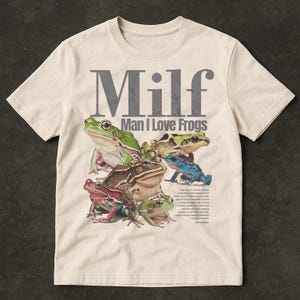 May include: Cream-coloured t-shirt featuring a graphic of various frogs in shades of green, brown, red, and blue. The text "Milf Man I Love Frogs" is printed above the frog illustration. The shirt has a crew neck and short sleeves.