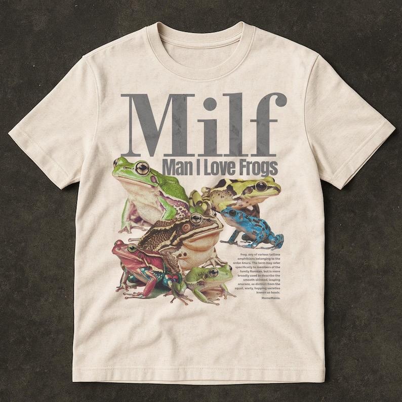 May include: Cream-coloured t-shirt featuring a graphic of various frogs in shades of green, brown, red, and blue. The text "Milf Man I Love Frogs" is printed above the frog illustration. The shirt has a crew neck and short sleeves.