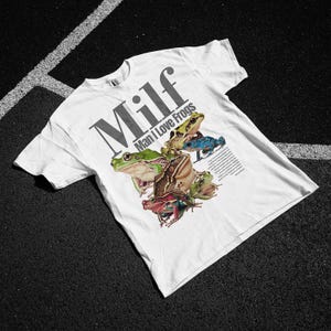 May include: White t-shirt with the word "Milf" in large grey letters above the phrase "Man I Love Frogs." The shirt features a colourful graphic of several frogs in various colours.