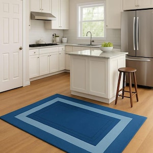May include: A rectangular blue rug with a lighter blue border and a darker blue center sits on a hardwood floor in a kitchen. The kitchen features white cabinets, stainless steel appliances, and a small island.