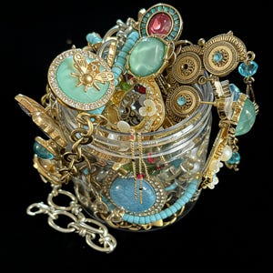 May include: A clear glass jar overflowing with an assortment of vintage jewelry. The jewelry includes gold-toned necklaces, bracelets, and rings, many adorned with turquoise, teal, and pink gemstones. Some pieces feature intricate floral and insect designs.