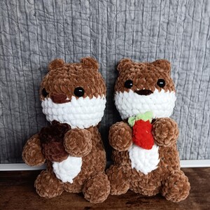 May include: Two handmade crochet otter plushies. The otters are brown with white chests and faces, and black eyes. One otter holds a brown object, the other holds a red strawberry with green leaves. The background is a gray quilted fabric.