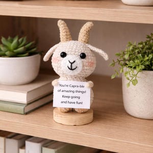 Puede incluir: Figura de cabra de ganchillo crema con cuernos y orejas beige y ojos negros. Sostiene un letrero con el texto "You're Capra-ble of amazing things! Keep going and have fun!". Sobre un soporte de madera.