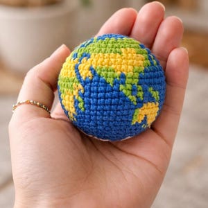 May include: A crocheted globe featuring blue oceans, green landmasses, and yellow continents. The handmade sphere is held in an open hand, showcasing the intricate stitchwork and vibrant colors. A small, gold ring adorns the finger.