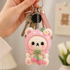 May include: A handmade crochet keychain featuring a white bear wearing a pink strawberry-themed hood. The bear has black eyes and a small black nose. The keychain includes a metal ring, a pink leather strap, and a chain holding keys.
