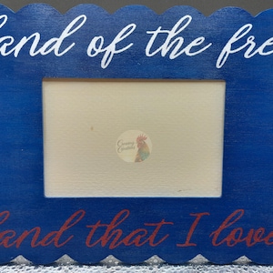 May include: A blue wooden picture frame with a scalloped edge. The frame has white script that reads "land of the free" at the top and red script that reads "land that I love" at the bottom. The frame has a rectangular opening.