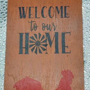 May include: Wooden door hanger with the words "WELCOME to our HOME" in black lettering. A red rooster silhouette is at the bottom. The hanger has a hole at the top for hanging.