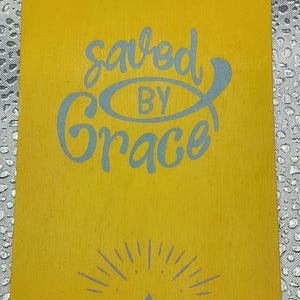 May include: Yellow door hanger with the words "saved BY Grace" in light blue script. Below the text is a mountain range and sun design in light blue. The words "HIS Grace" are at the bottom.