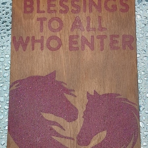 May include: Wooden door hanger with the text "BLESSINGS TO ALL WHO ENTER" in red. Features two glittery red horse head silhouettes. Includes a string for hanging and a small logo.