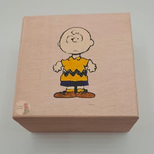 May include: A wooden box with a hand-painted image of Charlie Brown from the Peanuts comic strip. Charlie Brown is wearing his signature yellow shirt with a black zig-zag pattern and brown shoes. The box is light brown.