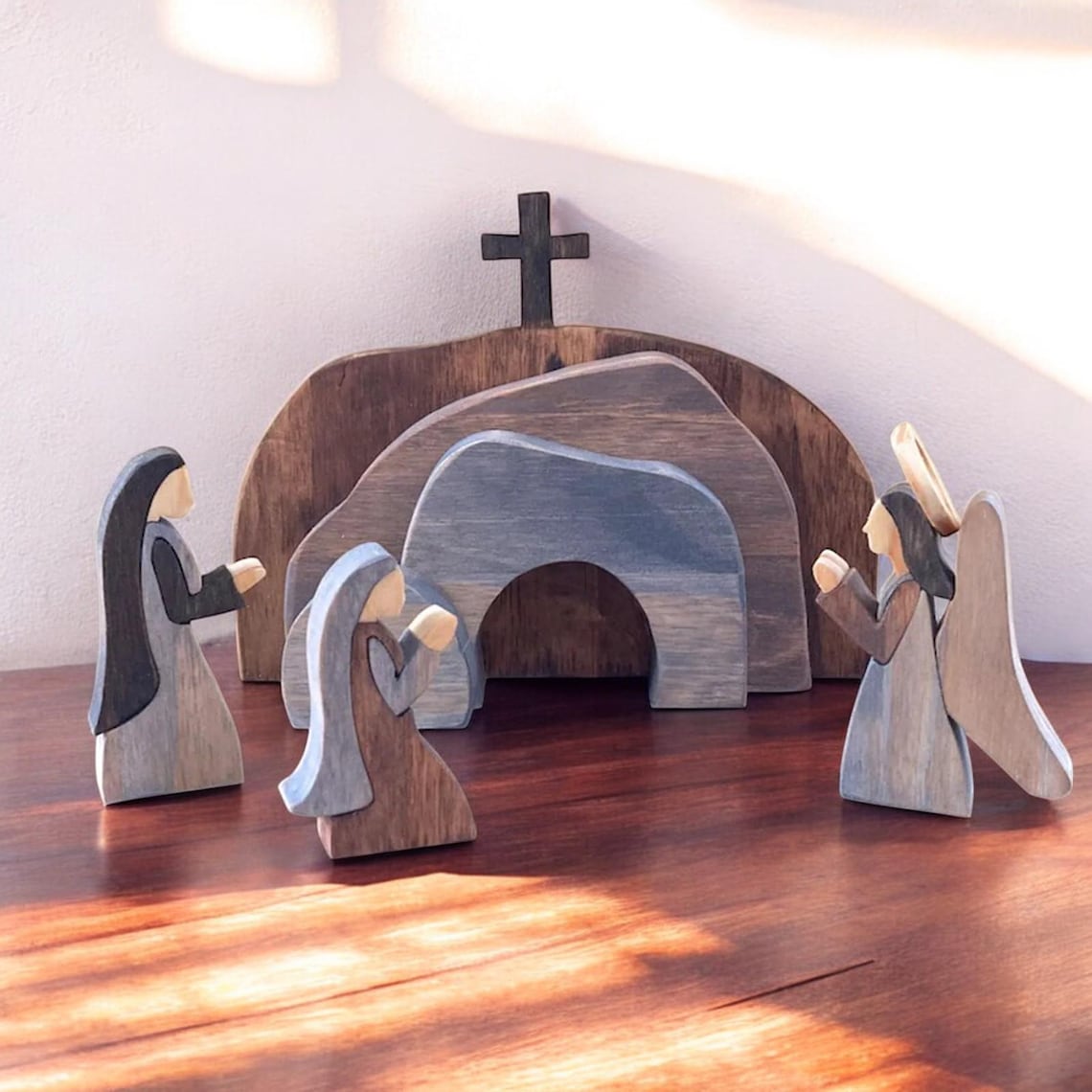 May include: Wooden figurines of three women and an angel standing in front of a wooden tomb with a cross on top. The tomb is made of three pieces of wood, each painted in a different shade of blue. The women are painted in shades of blue and brown. The angel is painted in shades of blue and gold. The figurines are on a wooden surface.