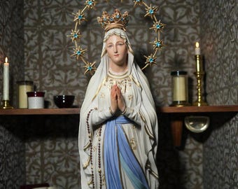 May include: A detailed statue of the Virgin Mary, wearing a crown and a halo of stars. She is dressed in a white robe with blue accents and a rosary. The statue is placed in a small alcove with candles and a book, creating a peaceful, religious setting.