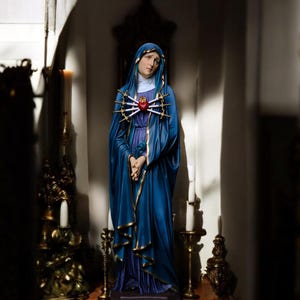May include: A statue of a woman in a long blue robe with gold trim, a white head covering, and a red heart pierced by swords on her chest. The statue is on a wooden surface, with candles and decorative objects in the background.